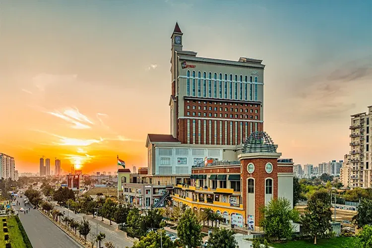 AIPL Joy Street high-rise building in Gurugram at golden-hour sunset, showing its twin clock towers, red-brick and white facade with tall arched glass windows, 