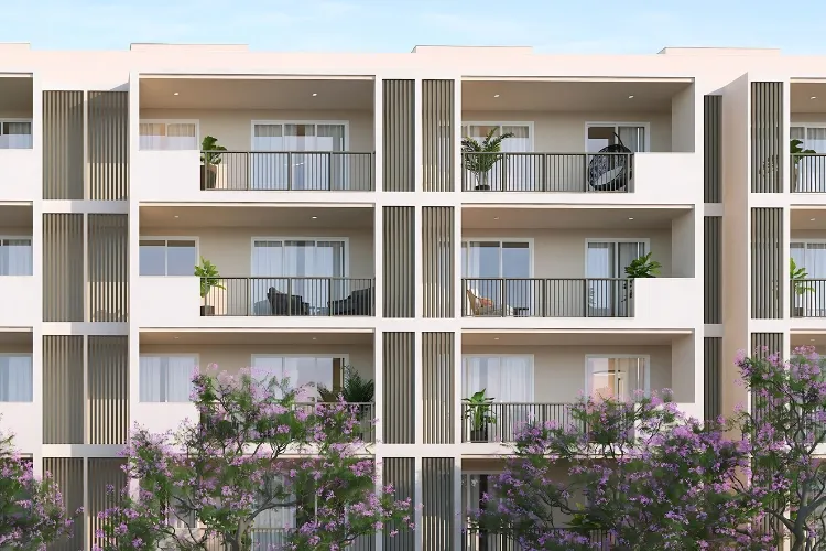 Close-up of a multi-story modern residential building facade at DLF Garden City, showing numerous balconies with dark railings, some featuring potted plants and outdoor seating, and purple flowering trees in the foreground.