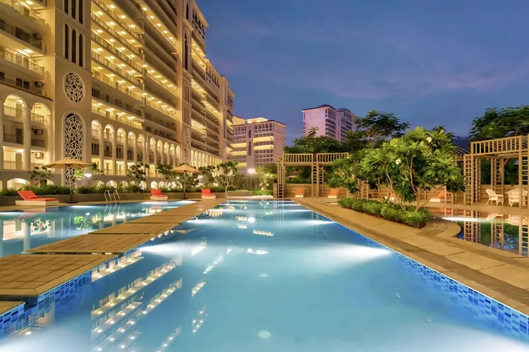 A beautifully lit swimming pool and surrounding deck area at DLF Skycourt at dusk, with the multi-story residential buildings illuminated in the background.
