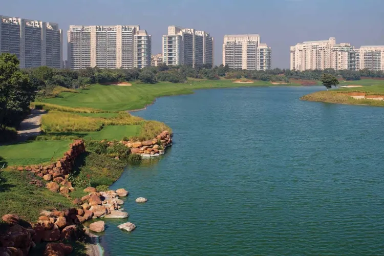 A wide outdoor shot featuring a large, calm body of blue-green water in the foreground, with lush green golf course fairways and a sandy bunker visible on the left. Several modern high-rise residential buildings with numerous balconies stand prominently in the background under a clear sky.