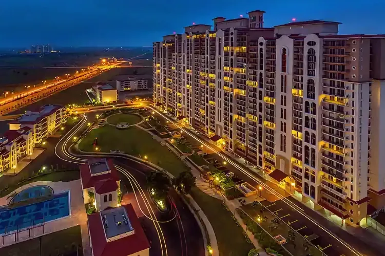 An aerial nighttime view of DLF The Magnolias residential complex on Golf Course Road, Gurgaon, showcasing multiple high-rise towers with illuminated balconies, meticulously landscaped grounds with winding pathways, and a swimming pool, all set against a dark sky with city lights.
