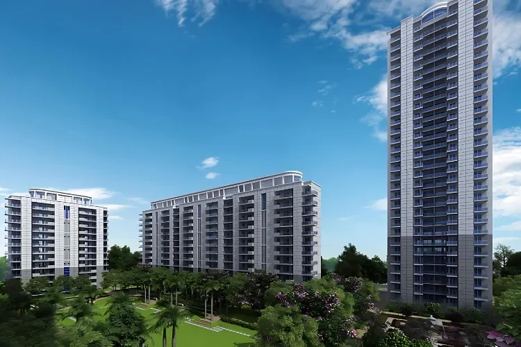 An exterior rendering of DLF The Ultima, showing three modern high-rise residential towers surrounded by meticulously landscaped green areas, trees, and pathways under a clear blue sky.