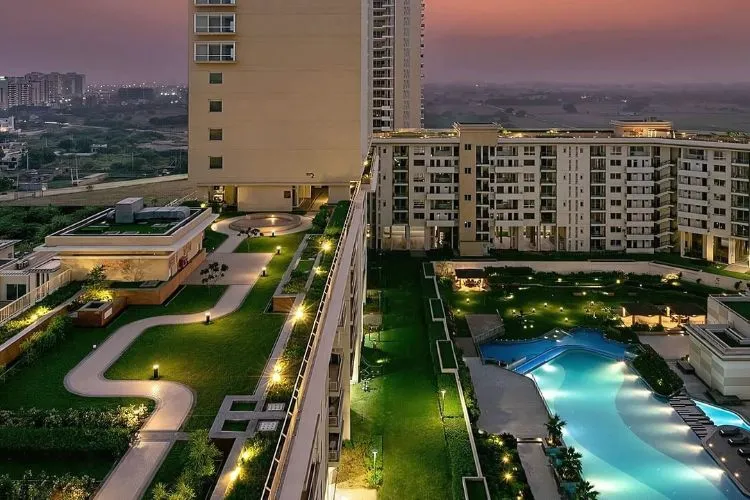 A beautiful twilight aerial photo of Experion Windchants, a residential complex. The image highlights the building's rooftop garden, which features lush green lawns, winding pathways, and elegant lighting. An outdoor swimming pool with glowing blue water is visible on a lower terrace, flanked by manicured green spaces. The scene is set against a soft, purple-hued sky and the distant skyline of a city.