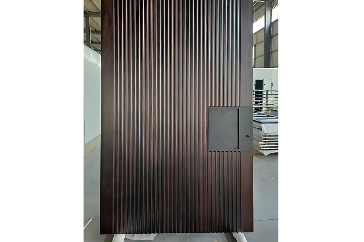  A close-up shot of a large, modern door made of a dark, reddish-brown fluted metal or wood. The vertical grooves cover the entire surface, except for a rectangular, matte gray section with a small handle positioned on the lower right side. The door appears to be in a warehouse or workshop setting, with an industrial background and stacked materials visible in the distance.