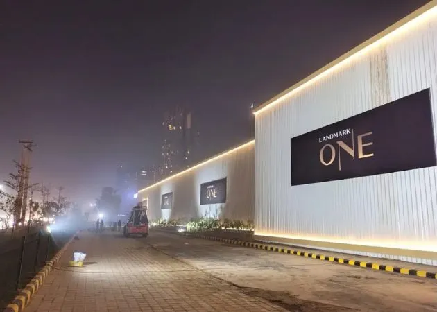 Landmark One Construction Site Boundary Wall at Night Nighttime view of the Landmark One construction site boundary wall with illuminated white paneling and a black signage board displaying the "LANDMARK ONE" logo in gold and white lettering, alongside a paved access road with yellow-and-black safety barriers.