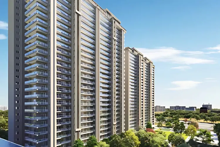 A sunny daytime rendering of Godrej Vrikshya residential towers in Gurgaon, showing a series of modern high-rise buildings with many balconies and large windows. The buildings are surrounded by green trees and landscaping, with a road visible in the foreground and other buildings in the distance against a clear blue sky.