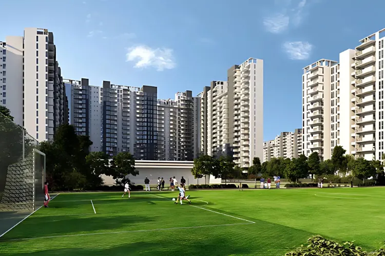 A wide-angle shot of a lush green football field at IREO The Corridors in Gurgaon. Several people are playing football, with others watching from the sidelines. In the background, modern, high-rise residential towers of the complex rise against a clear blue sky. The image highlights the on-site sports amenities and the expansive open spaces available for residents.