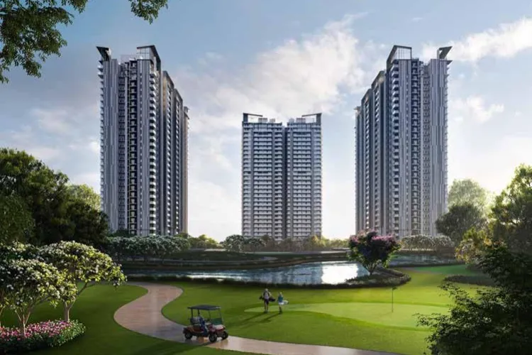 A panoramic view of three modern, tall residential towers of M3M Golf Hills, with balconies, set behind a lush green golf course featuring a winding path, a golf cart, a pond, and golfers, all under a clear sky.