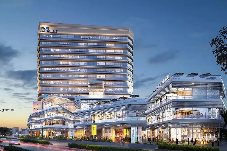 A modern, multi-story commercial building, M3M Paragon 57, featuring a towering office block above a multi-level retail complex with glass facades, illuminated at dusk. People are visible on the lower retail levels.
