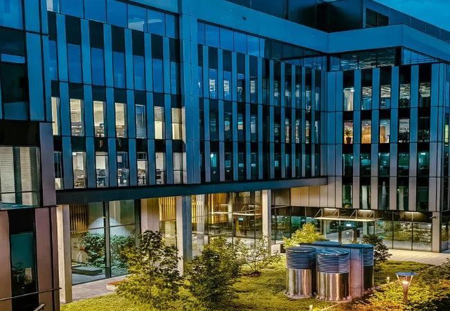 Night view of Emaar India Business Centre showing illuminated glass facade and landscaped inner courtyard with trees and modern metallic installations.