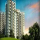 A tall, modern apartment building, Birla Pravaah in Sector 71, Gurgaon, stands against a vibrant sky with hues of blue and orange during sunset. Lush green grass and trees are in the foreground.